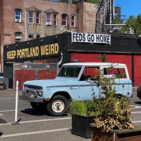 A sky blue Ford Bronco parked outside of Voodoo Doughnut in Portland Oregon with the Keep Portland Weird sign in the background.